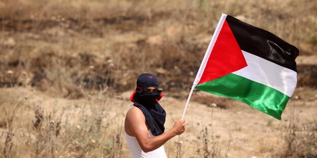Ein junger Mann hat sich ein T-Shirt um den Kopf gewickelt, er hält eine Palästina-Flagge