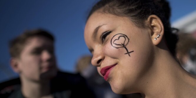 Eine junge Frau mit einem Frauen-Symbol auf der Wange, im Hintergrund verschwommen ein Mann