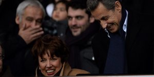 Roselyne Bachelot sitzt auf einer Tribüne, neben ihr steht Nicolas Sarkozy