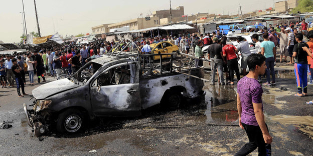 ein ausgebranntes Auto, im Hintergrund viele Menschen und Marktstände