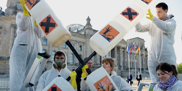 Menschen in weißen Overalls mit einem Kreuz aus Kanistern mit Gift-Warnhinweisen