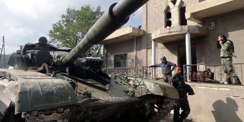 ein Panzer in Nahaufnahme, im Hintergrund ein zerstörtes Gebäude, davor einige Männer