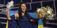 eine Frau hat einen Blumenstrauß und einen mikofonförmigen Pokal in den Händen, sie ist in eine ukrainische Fahne gehüllt