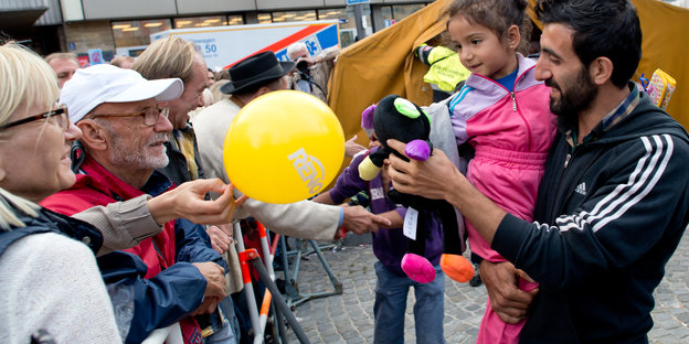 Einem Mann mit Kind auf dem Arm werden ein Ballon und ein Stofftier gereicht