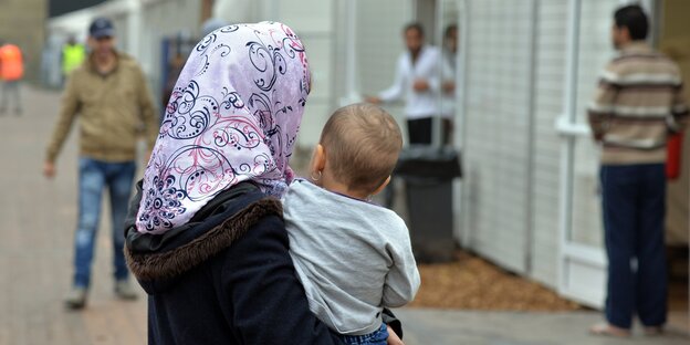 Eine Frau mit gemusterten Kopftuch steht mit Kind auf dem Arm vor weißen Container-Behausungen