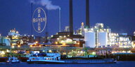 Abendhimmel und ein riesiges Bayer-Kreuz über einer Fabrik, im Vordergrund der Rhein