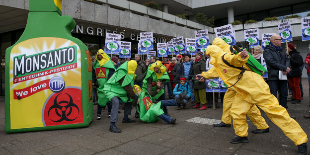 Zwei in gelbe Schutzanzüge gekleidete Umweltaktivisten besrpühen zu Boden gehende menschliche Maispflanzen. Im Hintergrund wird mit Plakaten gegen Glyphosat demonstriert.