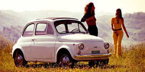 Eine junge Frau lehnt sich lässig an einen alten, weißen Fiat 500. Eine weitere kommt ihr entgegen