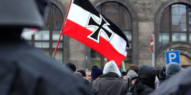 Hinter Polizisten stehen vermummte Rechte, eine Reichkriegsflagge mit einem Eisernen Kreuz weht darüber
