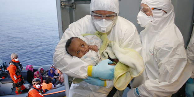 Zwei Marinesoldaten der Bundeswehr in weißen Schutzanzügen versorgen an Deck der Fregatte Hessen ein gerettetes Baby