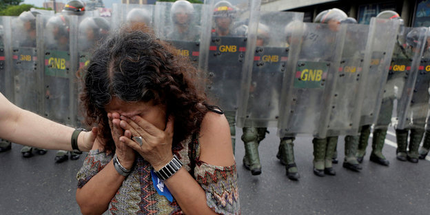 eine Frau hält sich die Hände vor das Gesicht, im Hintergrund Sicherheitskräfte mit großen Schilden