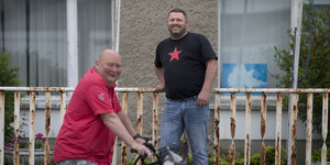 Zwei Männer, lachend vor einer Hausfassade. Einer der Männer sitzt auf einem Fahrrad