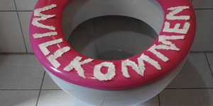 Ein Toilettenrand, auf den mit Buchstaben aus Klopapier das Wort "Willkommen" gelegt wurde