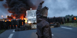 Ein Demonstrant hebt seine geballte Faust. Im Hintergrund sind ein Polizeieinsatzfahrzeug und Polizisten vor einem großen Feuer zu sehen
