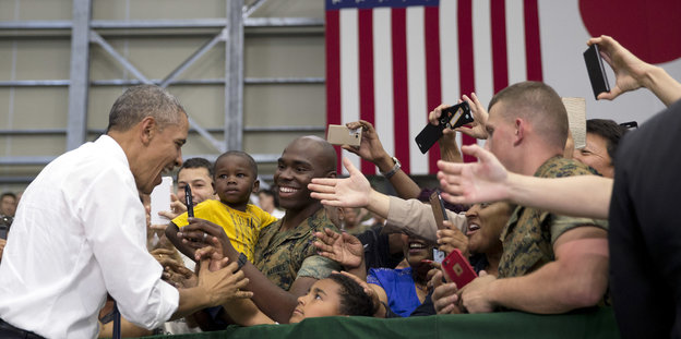 Obama begrüßt eine Gruppe von Menschen, im Hintergrund eine US- und eine Japan-Flagge