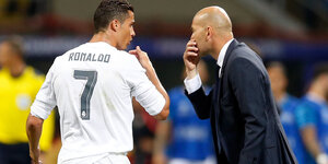 Zwei Männer (einer im Trikot, einer im Anzug) sprechen miteinander. Dabei halten sie sich jeweils eine Hand vor den Mund. Es sind Cristiano Ronaldo und Zinedine Zidane