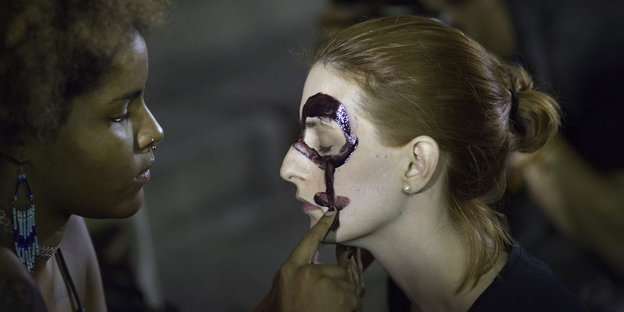 Eine Frau schminkt einer anderen Frau das Frauensymbol ins Gesicht.