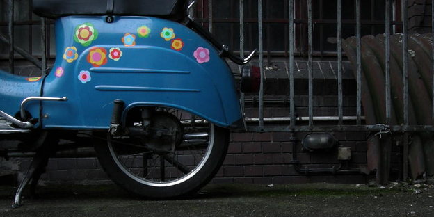 Ein blauer Motorroller mit Prilblumen beklebt steht an einem Zaun