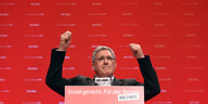 Bernd Riexinger von der Partei Die Linke steht mit erhobenen Armen an einem Rednerpult vor rotem Hintergrund