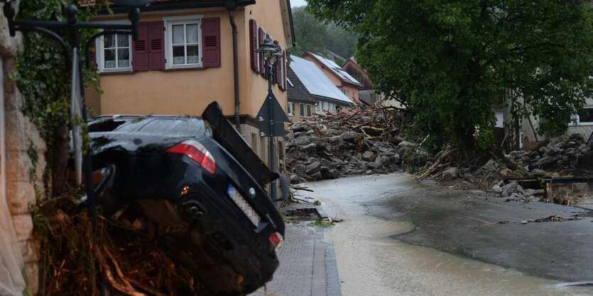 Ein Auto liegt quer auf einem Bürgersteig einer Dorfstraße, im Hintergrund Geröll