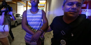 Ein Polizist führt einen Verdächtigen in Handschellen ab. Links: Ein Kameramann