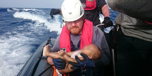 Ein Mann in Schwimmweste auf einem Schiff hält ein totes Baby im Arm