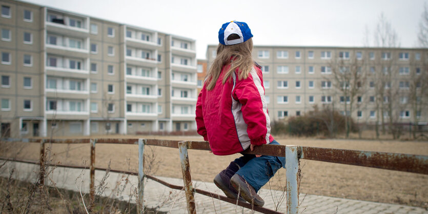 Ein Kind mit langen Haaren sitzt auf einem Zaun vor einem Plattenbau