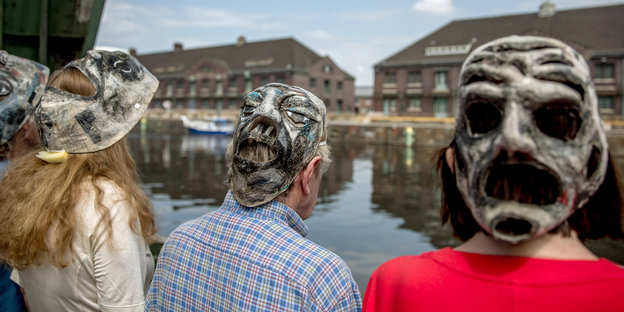 Gruselige Fratzenmasken auf den Hinterköpfen von Menschen, die vor einem Hafenbecken stehen