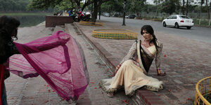 Eine Transgender-Person sitzt in einem rosafarbenen Kleid auf den Stufen vor einem Fluss, eine andere lässt einen lilafarenen Schal über ihrem Arm im Wind wehen