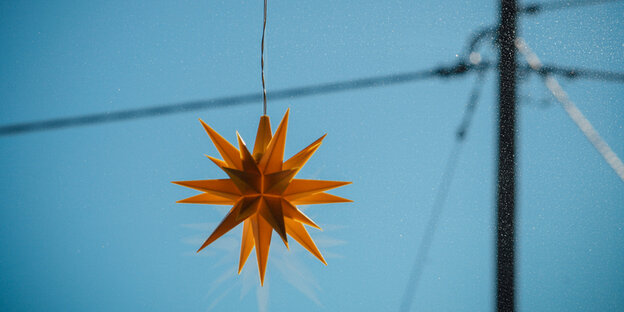 Ein gelber Stern hängt an einem Kabel. Im Hintergrund Teile eines Strommasts