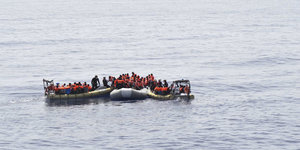 Auf dem Meer schwimmen mehrere Schlauchboote, in denen Menschen mit Rettungswesten stehen