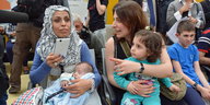 Zwei Frauen, die linke trägt ein Kopftuch, sitzen mit ihren kleinen Kindern auf dem Schoß in einem Flughafen