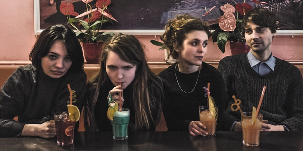Die Mitglieder von "Die Heiterkeit", drei Frauen, ein Mann, sitzen an einer Bar und trinken bunte Getränke