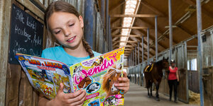 Ein Mädchen steht in einem Stall und liest ein buntes Magazin, im Hintergrund: ein Pferd