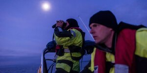 Zwei Männer mit Schwimmwesten in einem Boot auf dem Meer, einer schaut durch ein Fernglas