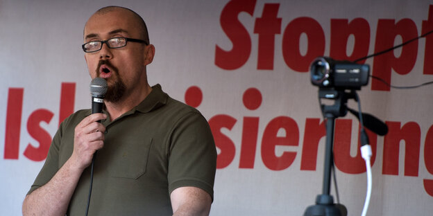 Ein Mann mit kurzen Haaren und Bart spricht in ein Mikrofon, im Hintergrund steht "Stoppt Islamisierung"