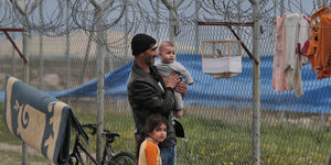 Ein Mann steht mit zwei Kindern vor einem Stacheldrahtzaun, an dem ein winziger Vogelkäfig hängt