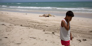 Ein Junge am Strand, dahinter unter einer Decke eine Leiche