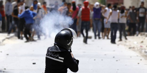 Ein Polizist mit Helm und arabischer Schrift auf der Jacke ist von hinten zu sehen, er feuert in Richtung einer Menschenmenge