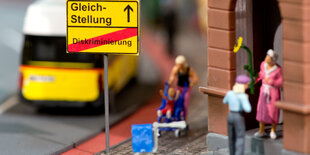 In einem Playmobildiorama sind zwei Frauen zu sehen, eine hält eine Blumen in den Händen, daneben steht ein Schild mit der Aufschrift „Gleichberechtigung“, darunter durchgestrichen „Diskriminierung“