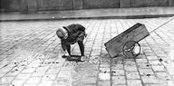 Schwarz-weiß-Fotografie: Ein kleiner Junge schiebt Kohlenstücken auf eine Schippe, neben sich ein Ziehwägelchen