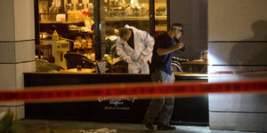 Zwei Männer untersuchen ein Restaurant, in dem ein Anschlag stattgefunden hat. Davor sieht man ein Absperrband