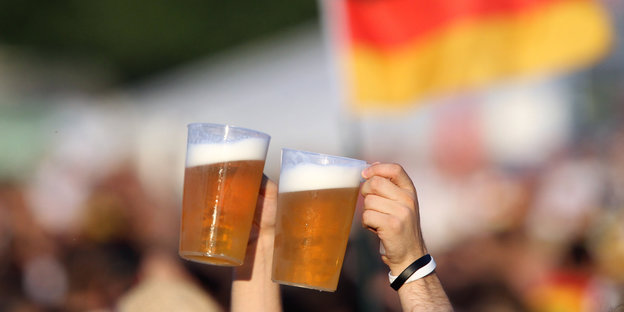 Zwei Bierbecher werden hochgehalten, im Hintergrund eine Menschenmenge und eine Deutschlandfahne