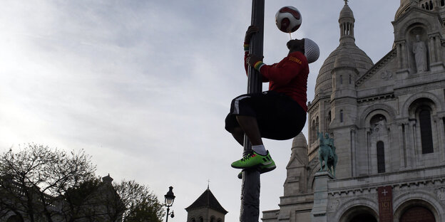 Ein Mann klettert eine Laterne hoch und jongliert mit einem Fußball