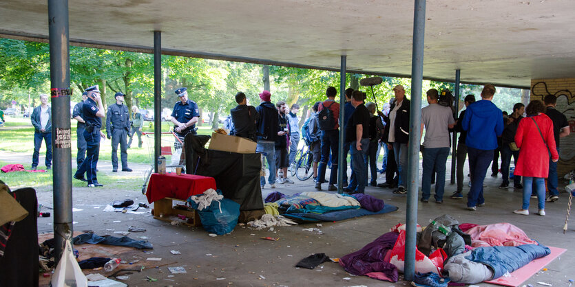 Unter einem Vordach liegen verstreut die Habseligkeiten von Menschen. Im Hintergrund stehen Polizisten.