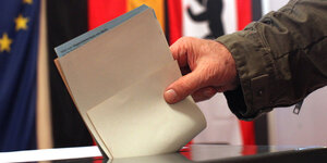 Eine Hand wirft Zettel in eine Wahlurne, im Hintergrund die Fahnen Berlins, Europas und Deutschlands