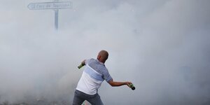 Tränengas vernebelt eine Straße. Ein glatzköpfiger Mann schmeißt Flaschen in die Wolke.