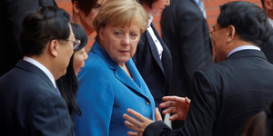 Angela Merkel, eine Frau kurzen hellroten Haaren und blauem Blazer, mit einer Gruppe chinesischen Frauen und Männern.