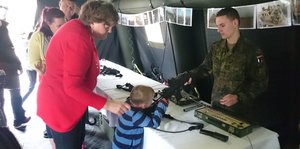 ein Kind mit einer Frau an einem Stand der Bundeswehr, das Kind hält ein Gewehr in der Hand, hinter dem Stand ein Mann in Uniform