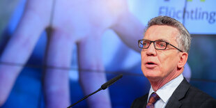 De Maizière redet, im Hintergrund eine Projektion mit einer Hand, auf der "WIR HELFEN" steht